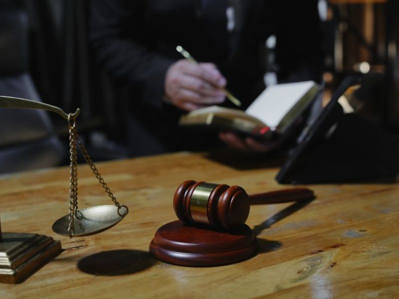 A wooden desk displays a gavel and a balance scale. A person, partially visible, writes in a notebook, indicating activity in a law-related context with soft lighting in the background.