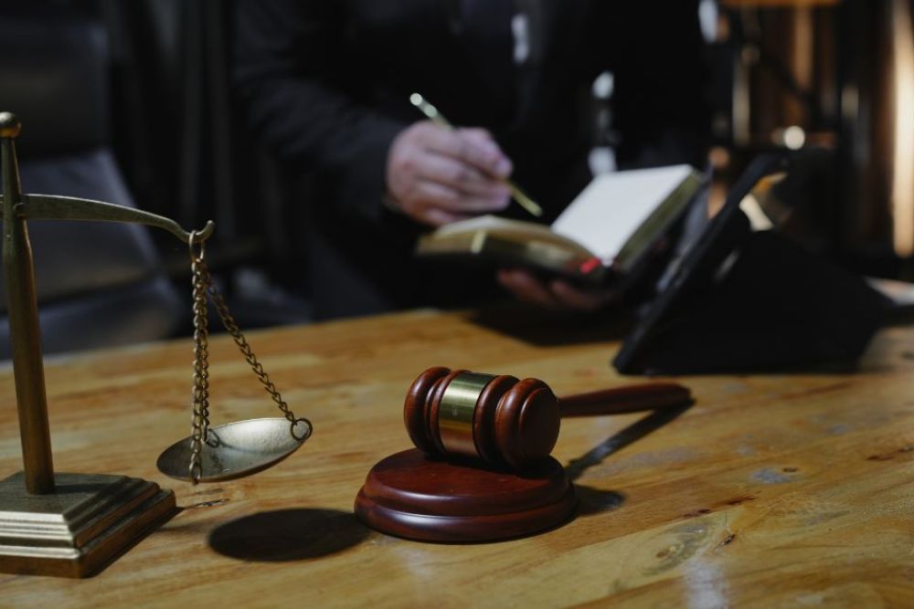 A wooden desk displays a gavel and a balance scale. A person, partially visible, writes in a notebook, indicating activity in a law-related context with soft lighting in the background.