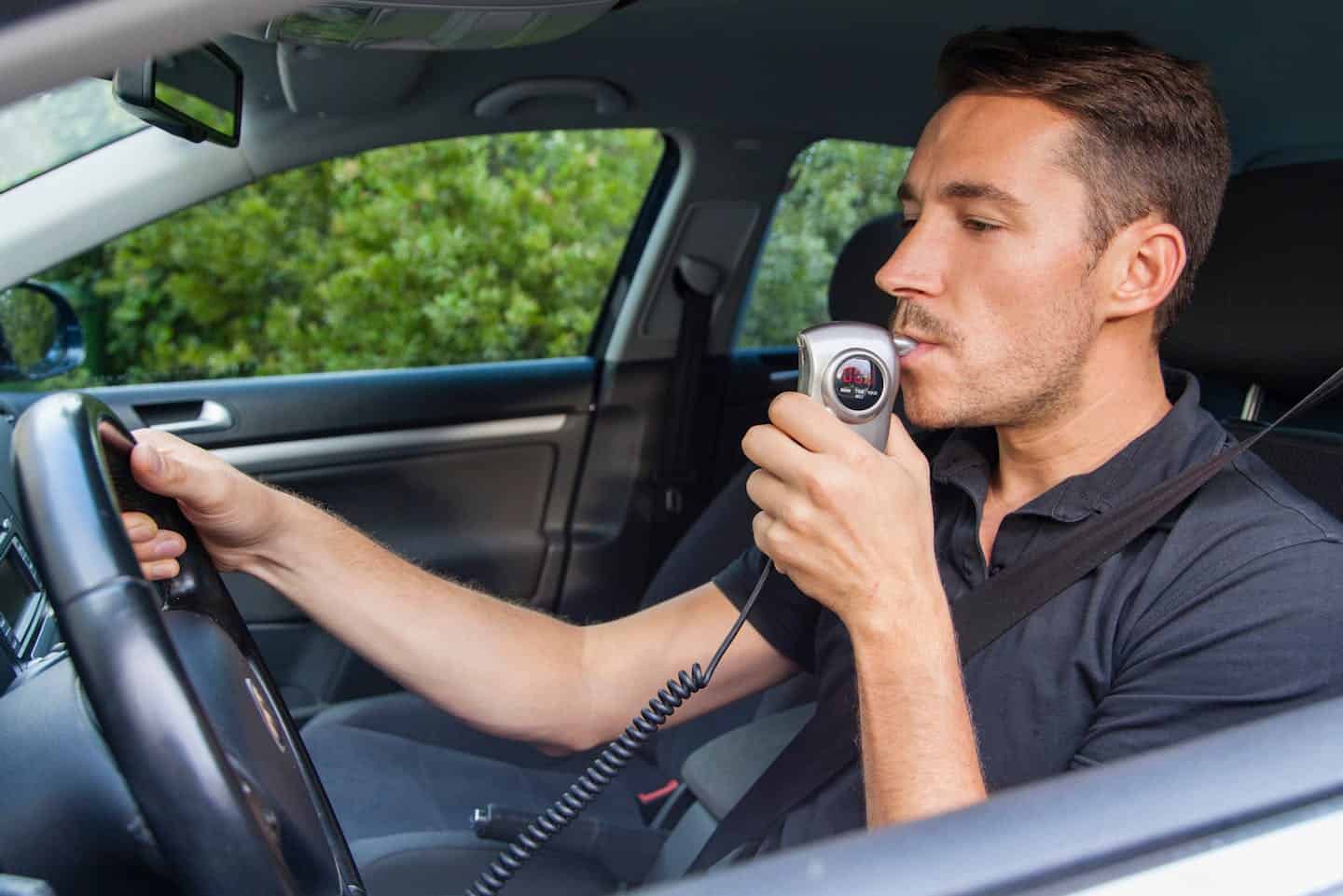 man blowing in BAC monitor attached to car with hand on steering wheel