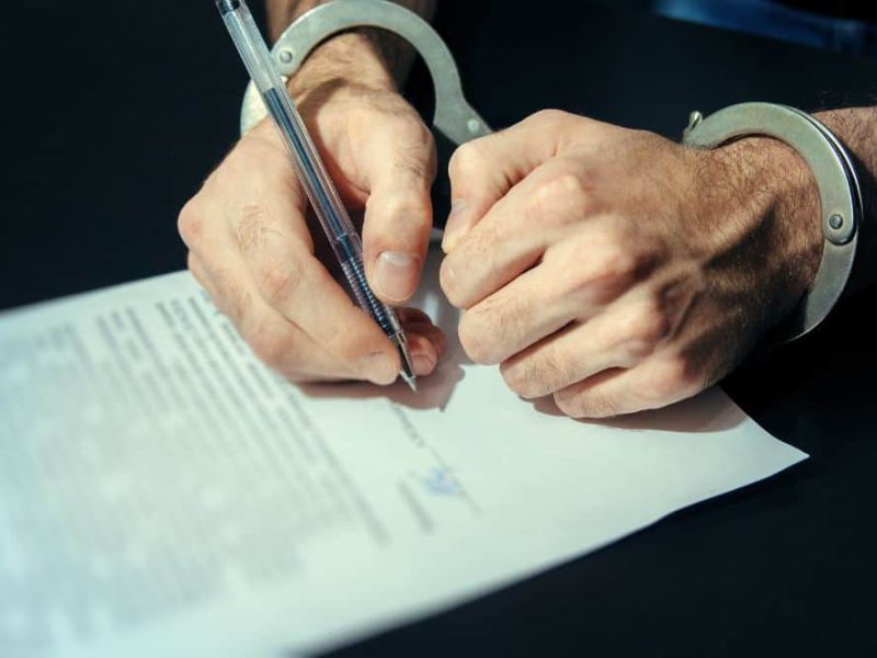 Man in handcuffs signing a plea bargain deal on a dark table with a silver pen