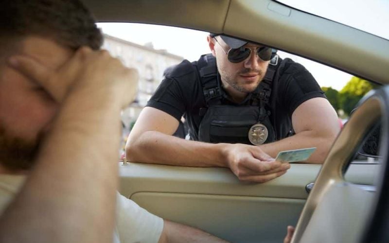 A police officer, wearing sunglasses and a badge, examines an drivers license while leaning into a car window. The driver, seated, appears distressed, resting his head on a hand.