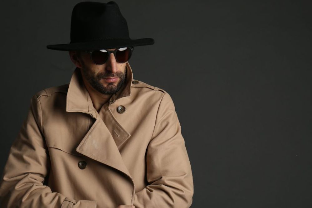 A man wearing a beige trench coat and a black hat poses confidently while adjusting his sunglasses, set against a dark, neutral background.