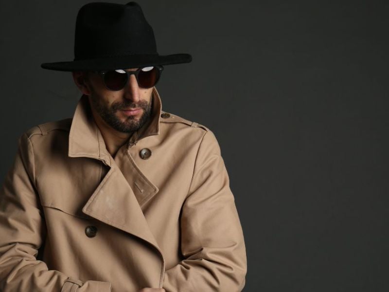 A man wearing a beige trench coat and a black hat poses confidently while adjusting his sunglasses, set against a dark, neutral background.