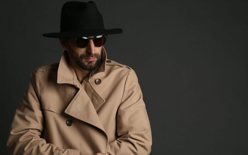 A man wearing a beige trench coat and a black hat poses confidently while adjusting his sunglasses, set against a dark, neutral background.
