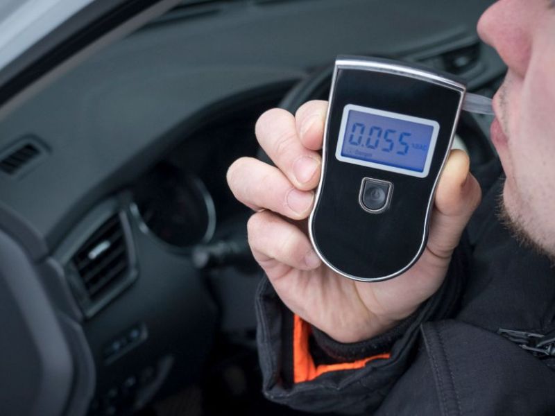 A person holds a breathalyzer to their mouth, exhaling into it. The device displays a reading of 0.055. The setting appears to be inside a vehicle.