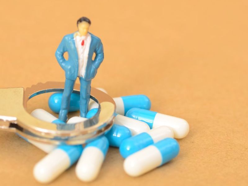 A miniature figure in a blue suit stands inside a silver handcuff, surrounded by various blue and white capsules. The scene rests on a brown background.