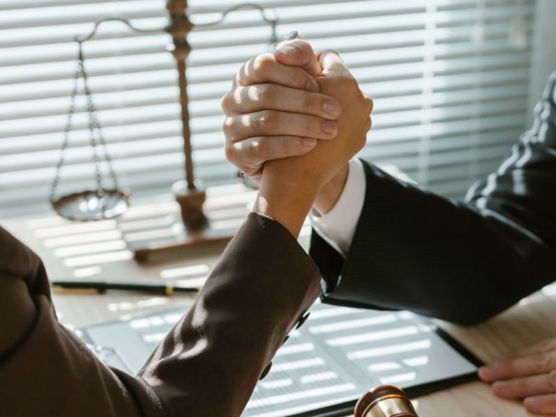 Two individuals engage in an arm wrestling match at a desk. A gavel and documents are visible nearby, with sunlight filtering through blinds, casting shadows in the office setting.