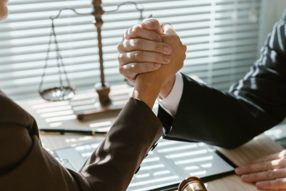 Two individuals engage in an arm wrestling match at a desk. A gavel and documents are visible nearby, with sunlight filtering through blinds, casting shadows in the office setting.