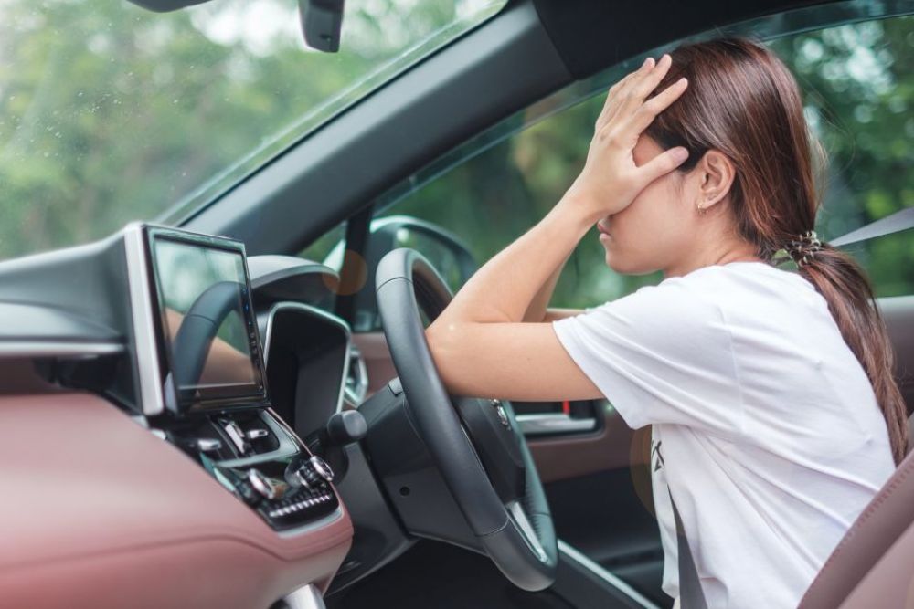A driver sits in a car, visibly distressed, with one hand on their forehead, surrounded by a modern dashboard and soft, natural light filtering through the windows.