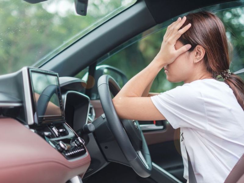 A driver sits in a car, visibly distressed, with one hand on their forehead, surrounded by a modern dashboard and soft, natural light filtering through the windows.