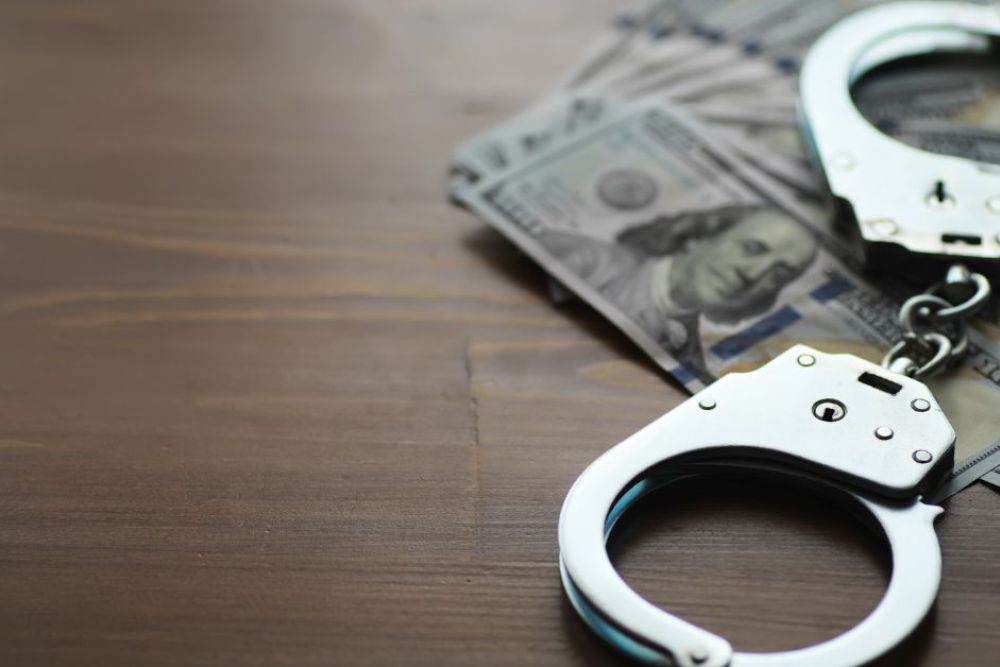 Handcuffs rest on a stack of hundred-dollar bills on a wooden surface, suggesting a connection between crime and money within a quiet, subdued environment.