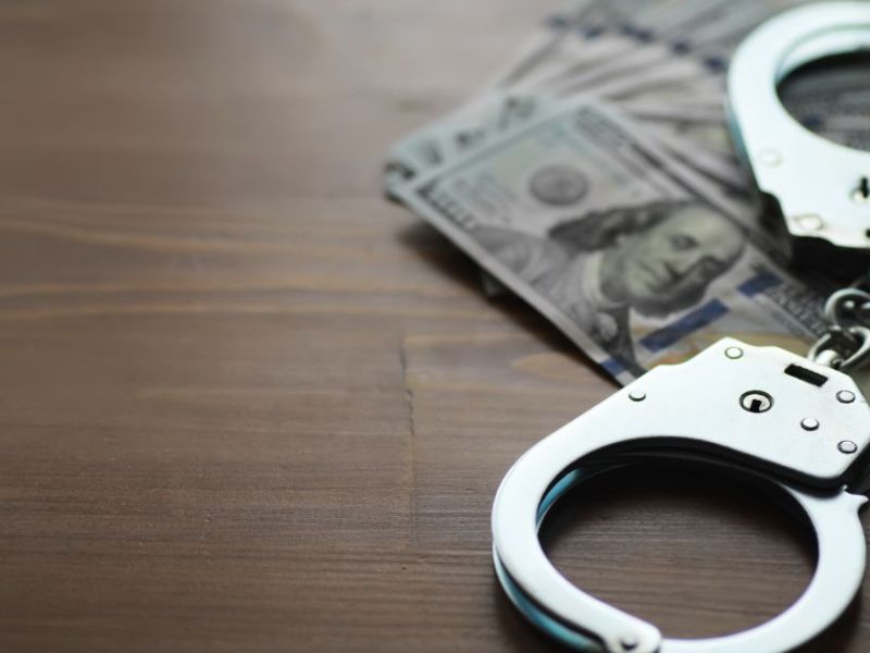 Handcuffs rest on a stack of hundred-dollar bills on a wooden surface, suggesting a connection between crime and money within a quiet, subdued environment.