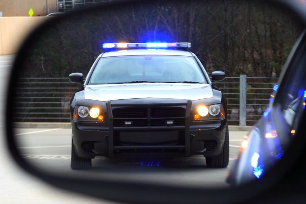 A police car with flashing blue lights is parked in front of another vehicle, visible in the side mirror. The setting is a parking area with trees in the background.