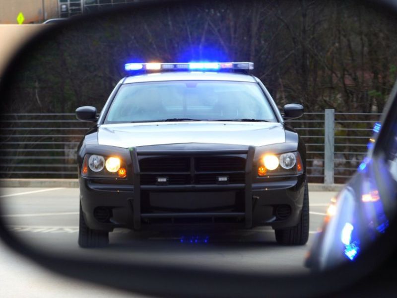 A police car with flashing blue lights is parked in front of another vehicle, visible in the side mirror. The setting is a parking area with trees in the background.