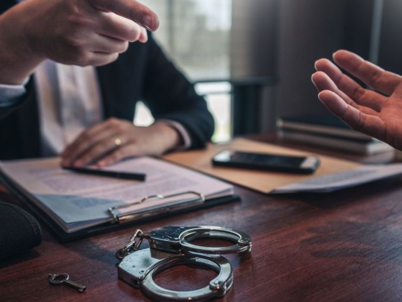Handcuffs lie on a wooden table while one hand gestures assertively towards another person, indicating a conversation in a formal setting, possibly a legal discussion or negotiation.