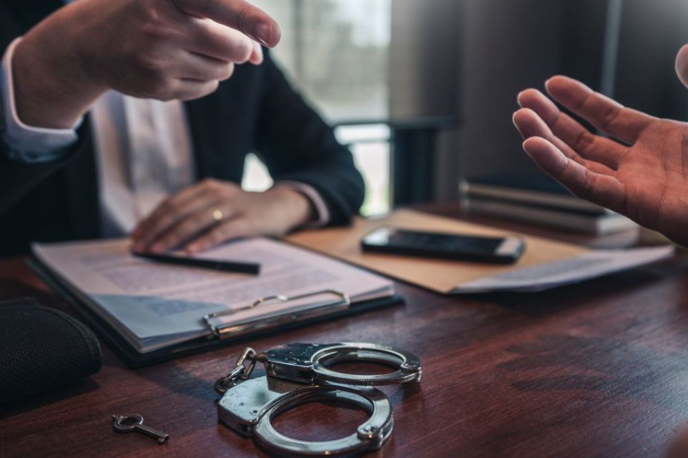 Handcuffs lie on a wooden table while one hand gestures assertively towards another person, indicating a conversation in a formal setting, possibly a legal discussion or negotiation.