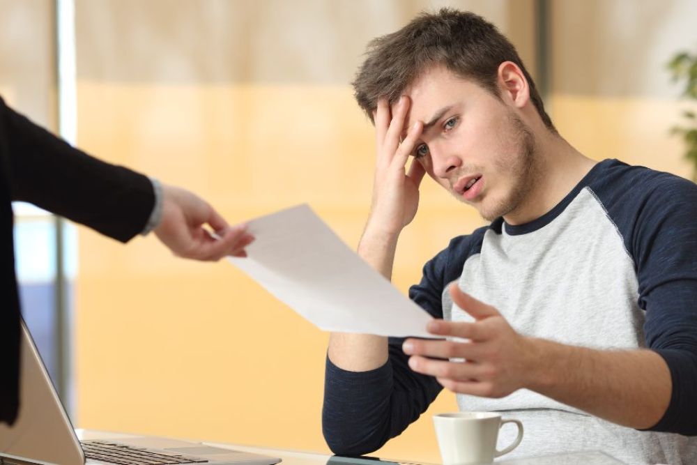 A young man sits at a desk, looking stressed while holding a paper with one hand and touching his forehead with the other. A person in a suit hands him the paper.