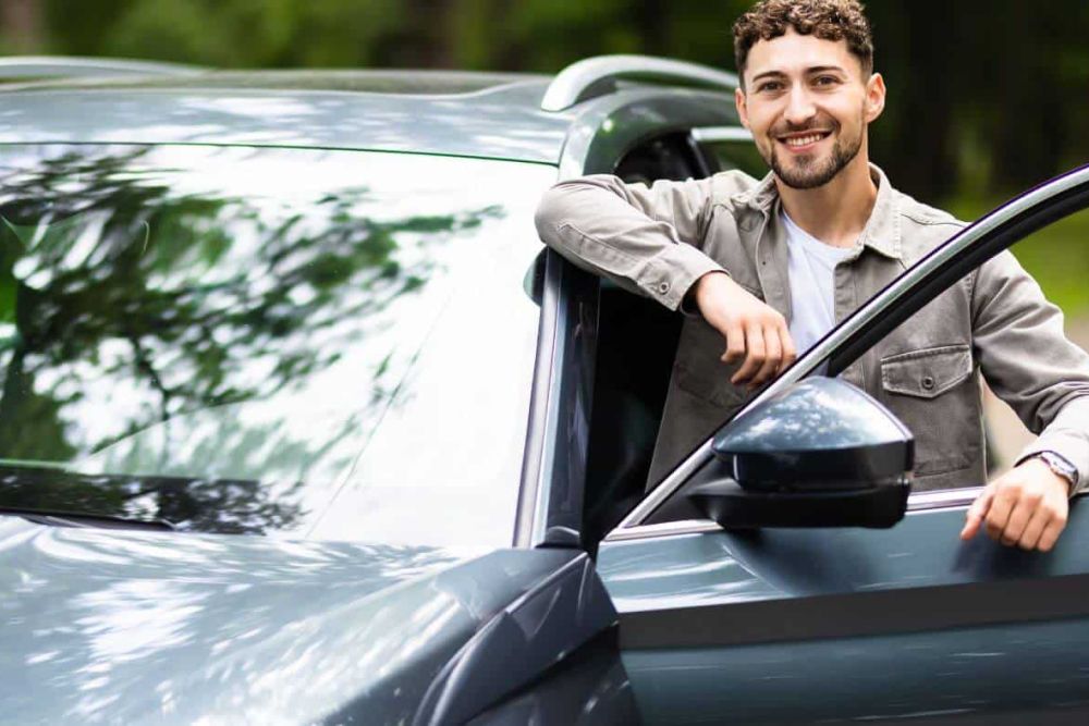 A smiling man leaning on an open car door, resting in a forested area.