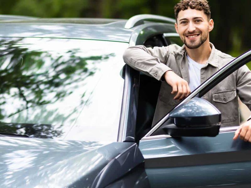 A smiling man leaning on an open car door, resting in a forested area.