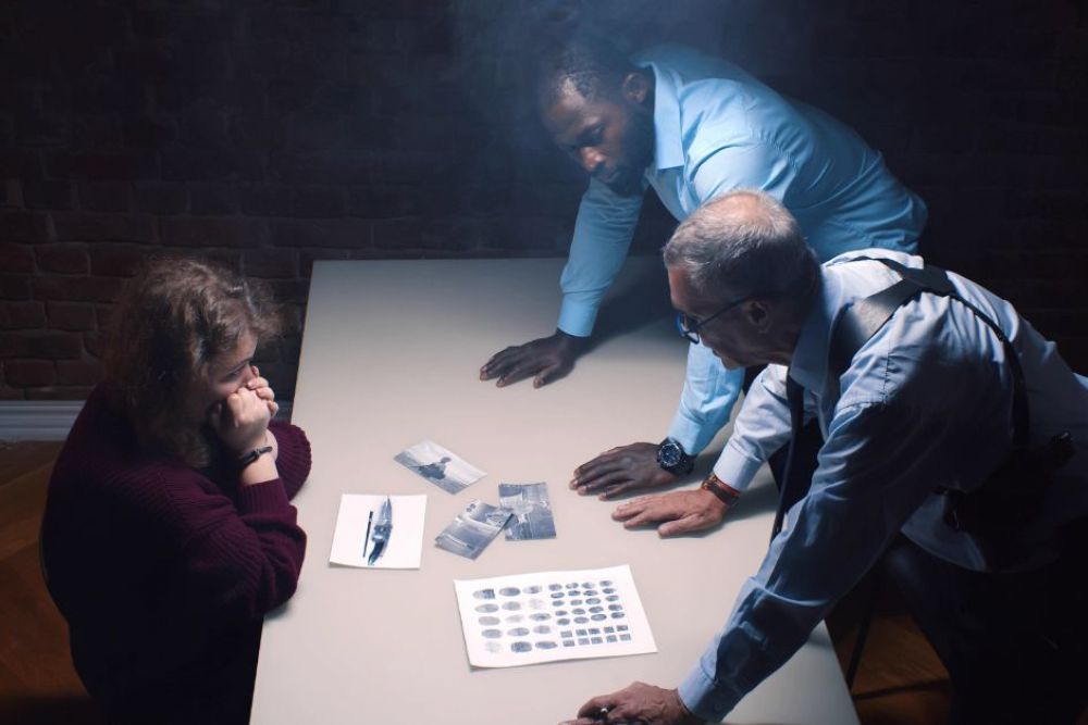 A woman sits anxiously at a table, while two men, one in a light blue shirt and the other in a suit, examine various photographs and papers under low lighting in a brick-walled room.