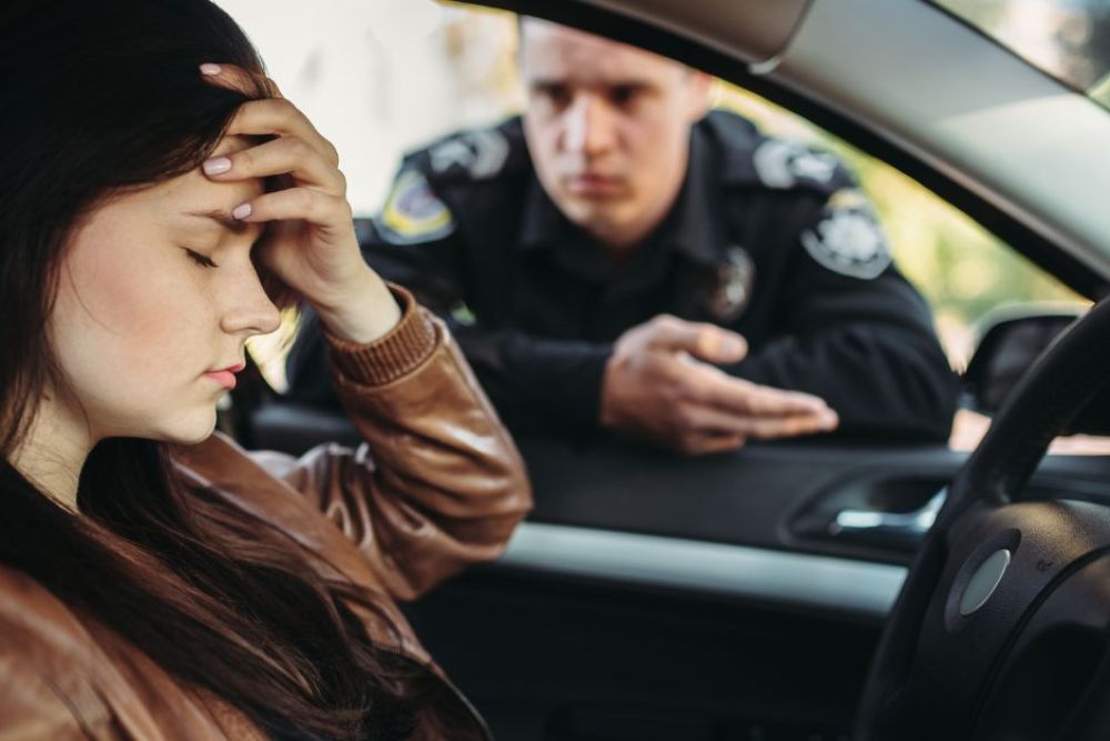 A young woman in a brown leather jacket rests her forehead on her hand, appearing distressed. A police officer leans into the car, gesturing and speaking to her.