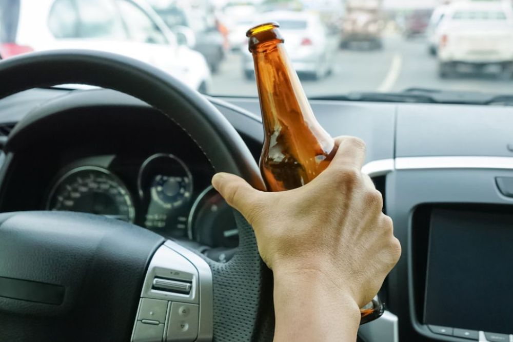 A hand grasping a brown bottle is positioned above a steering wheel, while the car is in traffic, surrounded by vehicles in a blurred urban environment.