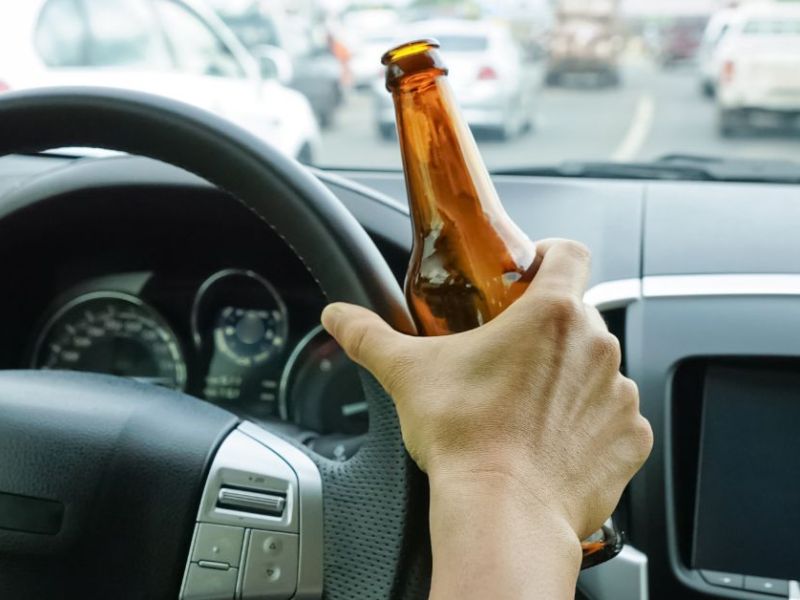 A hand grasping a brown bottle is positioned above a steering wheel, while the car is in traffic, surrounded by vehicles in a blurred urban environment.