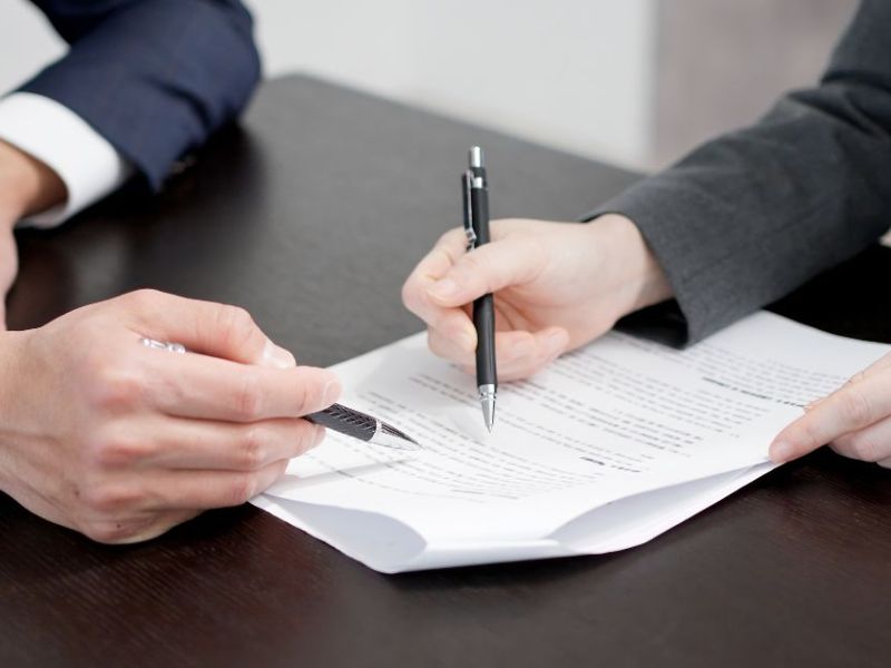 A hand holding a pen signs a document on a wooden table, while another hand points at the text. The setting appears to be a professional office environment.