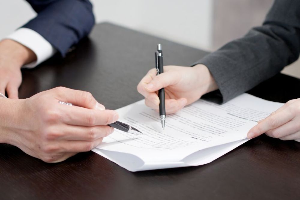A hand holding a pen signs a document on a wooden table, while another hand points at the text. The setting appears to be a professional office environment.