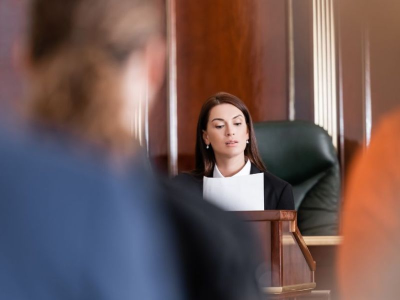 A judge, seated at a wooden bench, reads from a document. The courtroom setting features polished wood paneling and a green chair, with blurred figures of observers in the foreground.