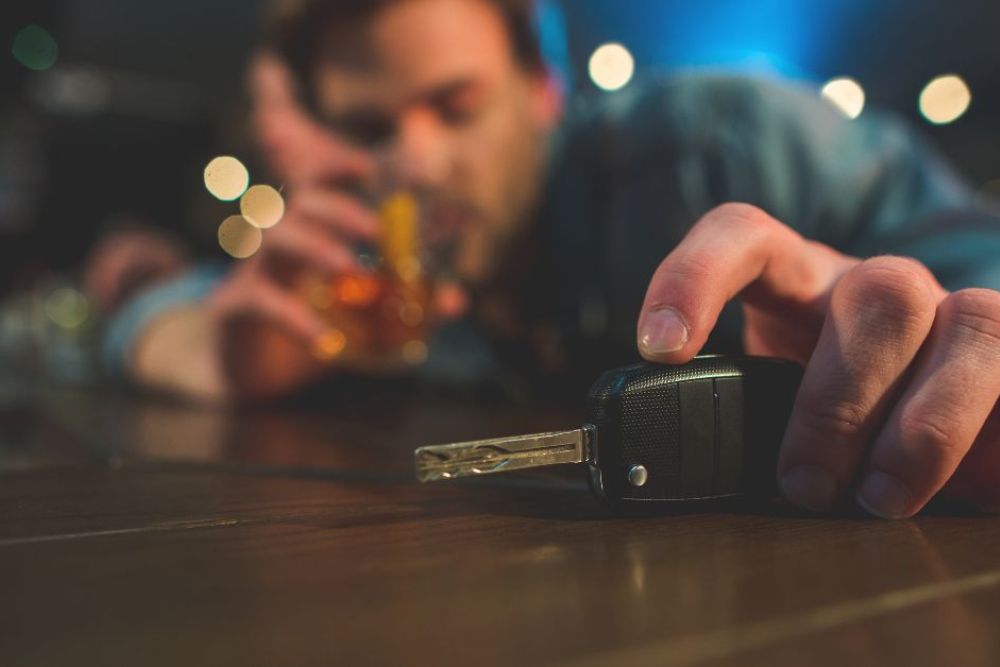 A car key lies on a wooden table while a person, blurred in the background, reaches for a drink. Dim lighting creates a relaxed bar atmosphere.
