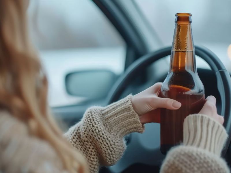 A person holds a bottle while driving, gripping the steering wheel with one hand. The environment suggests a vehicle interior, with blurred lights in the background indicating movement.