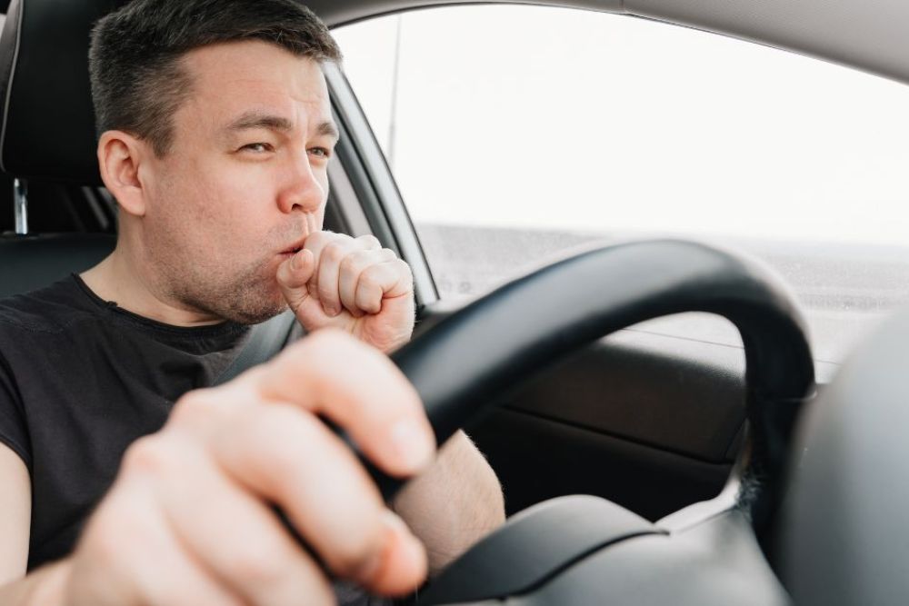 A man is driving a car, focused and coughing into his hand while gripping the steering wheel. The car's interior is visible, with a blurred outdoor landscape through the windows.