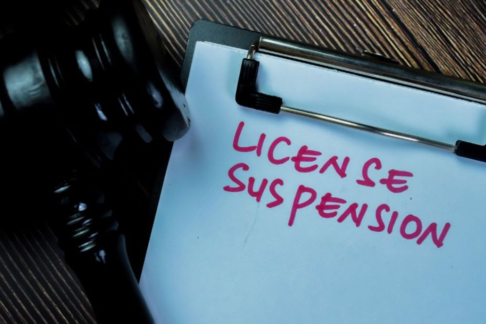 A gavel rests beside a clipboard displaying the words "LICENSE SUSPENSION" in bold, pink handwriting, set against a dark wooden surface, suggesting a legal context.