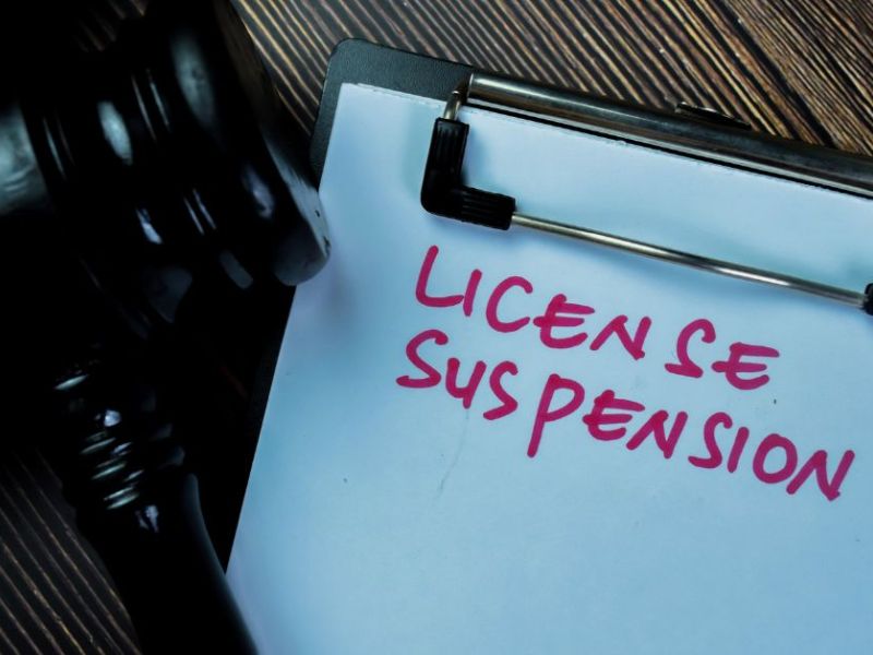 A gavel rests beside a clipboard displaying the words "LICENSE SUSPENSION" in bold, pink handwriting, set against a dark wooden surface, suggesting a legal context.