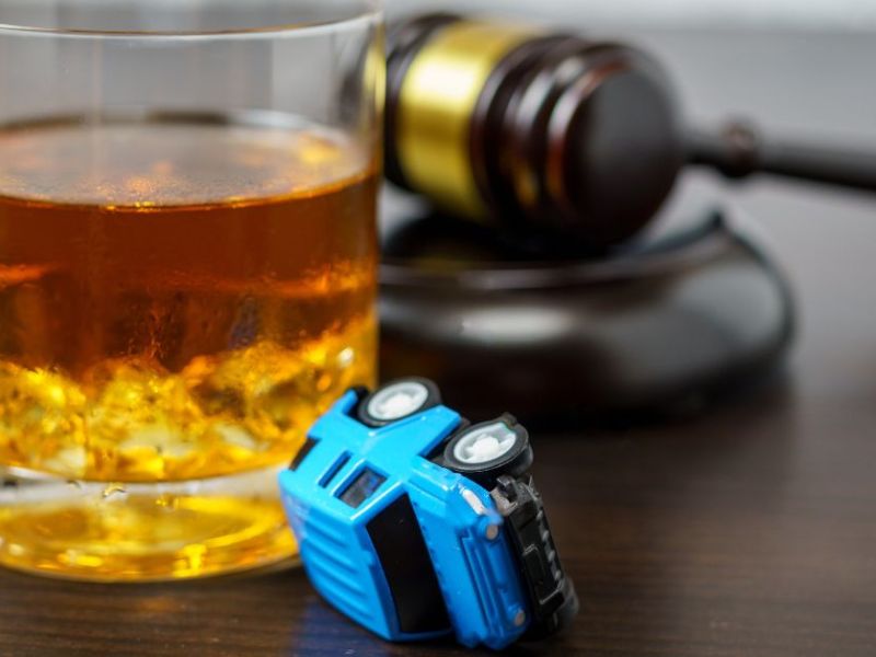 A glass of amber liquid with ice sits beside a tipped-over blue toy truck and a wooden gavel on a dark wooden surface, suggesting themes of judgment and playfulness.