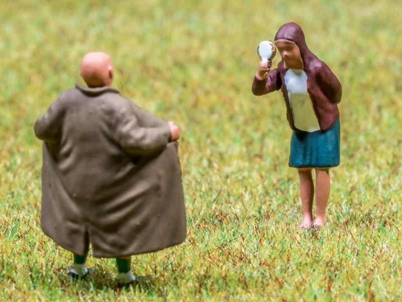 A small, stylized figure of a woman examines herself in a mirror, while a bald man in a coat stands with his back to her, on a grassy surface.