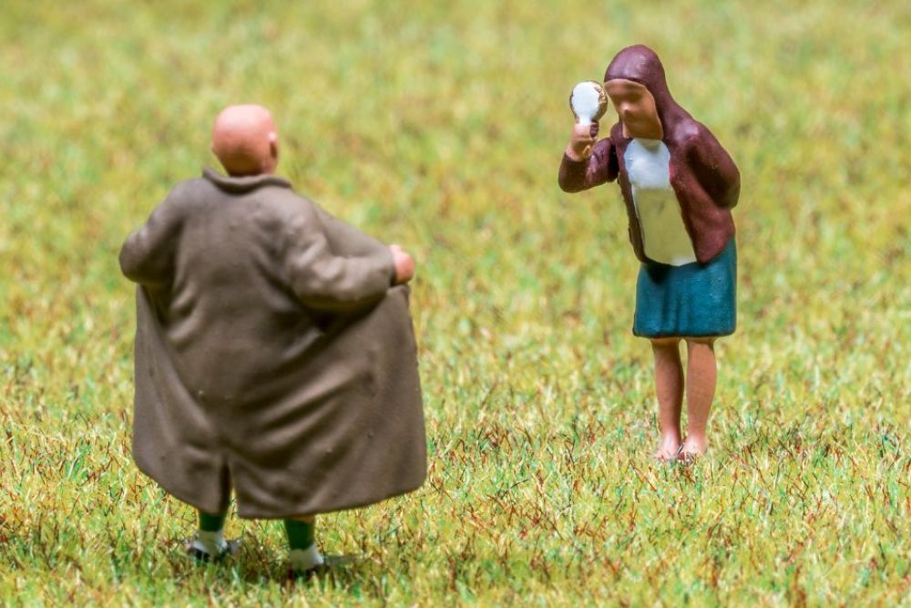 A small, stylized figure of a woman examines herself in a mirror, while a bald man in a coat stands with his back to her, on a grassy surface.