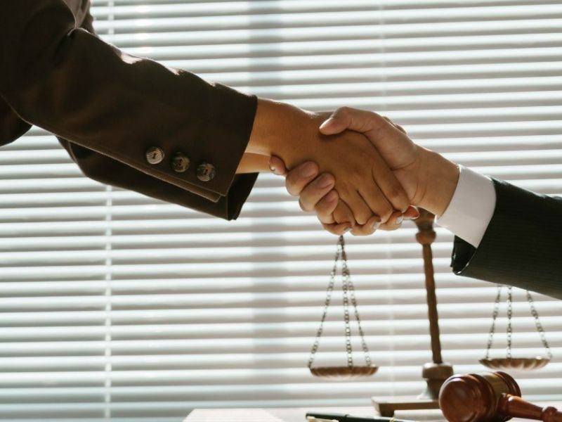 Two hands shake in a formal setting, suggesting agreement or partnership. In the background, a row of blinds allows light to filter in, with a scale of justice visible nearby.