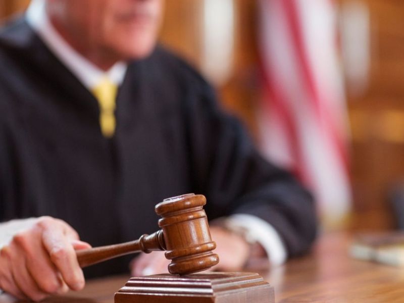A judge, wearing a black robe, strikes a wooden gavel on a desk. The courtroom features wooden paneling and an American flag in the background, indicating a formal legal setting.