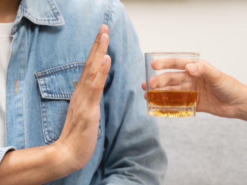 A person extends their hand to refuse a glass of amber liquid offered by another hand, set against a neutral-colored background, suggesting a conversation about drinking choices.
