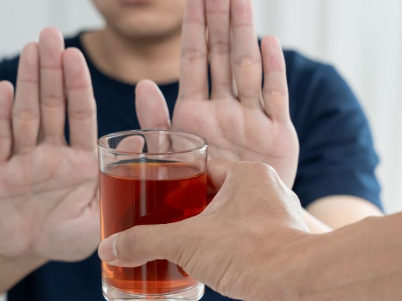 A person holds a glass of reddish liquid towards another individual, who raises both hands defensively in refusal, set in a bright, minimalist indoor environment.