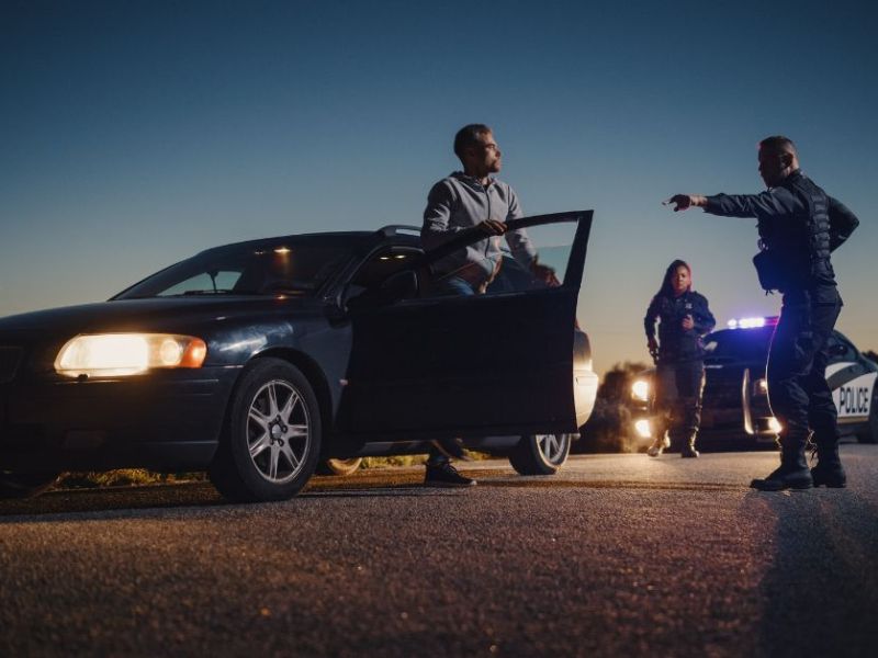A man stands outside a dark car with an open door, while two police officers engage with him on a deserted road at dusk, illuminated by police lights.