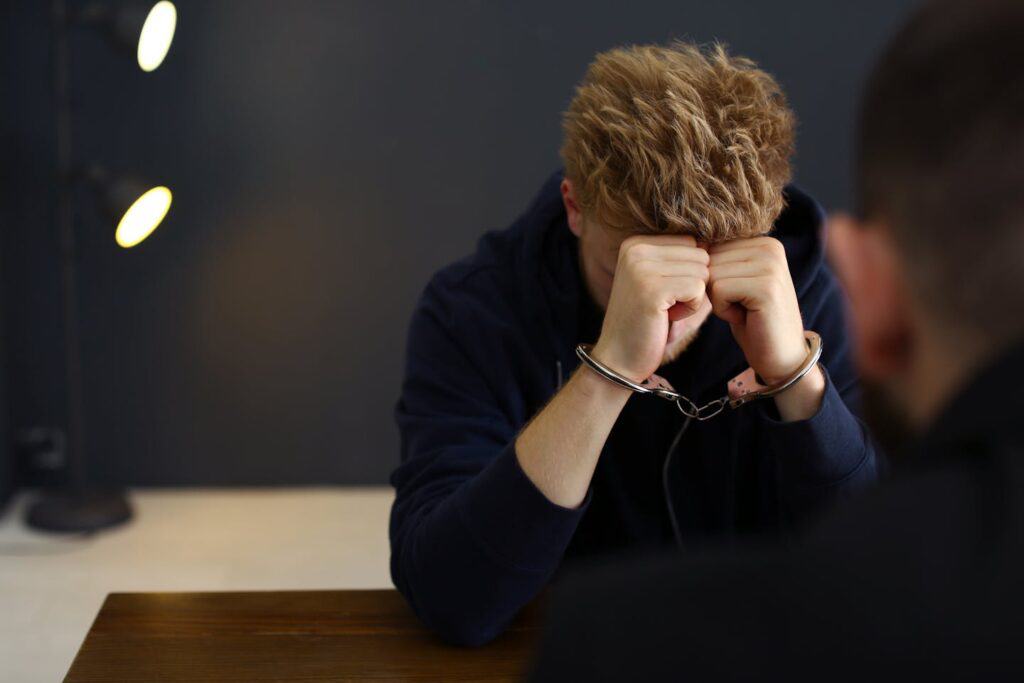 A young man, handcuffed, bows his head with hands clasped over his face, expressing distress. He sits at a wooden table in a dimly lit room, with a shadowy figure in the background.