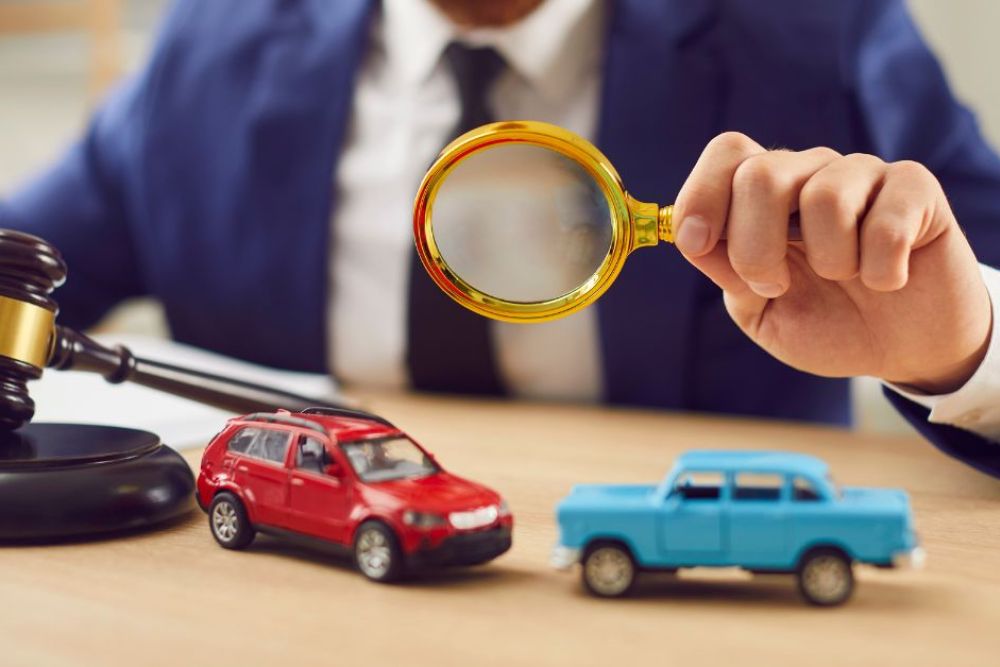 A hand holds a magnifying glass over two toy cars—a red SUV and a blue vintage car—on a desk near a gavel, suggesting examination or investigation in a legal context.
