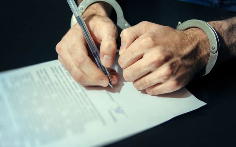 Man in handcuffs signing a plea bargain deal on a dark table with a silver pen