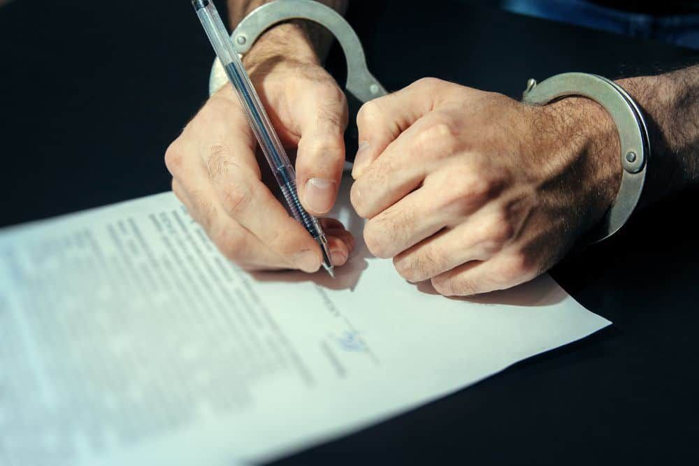 Man in handcuffs signing a plea bargain deal on a dark table with a silver pen
