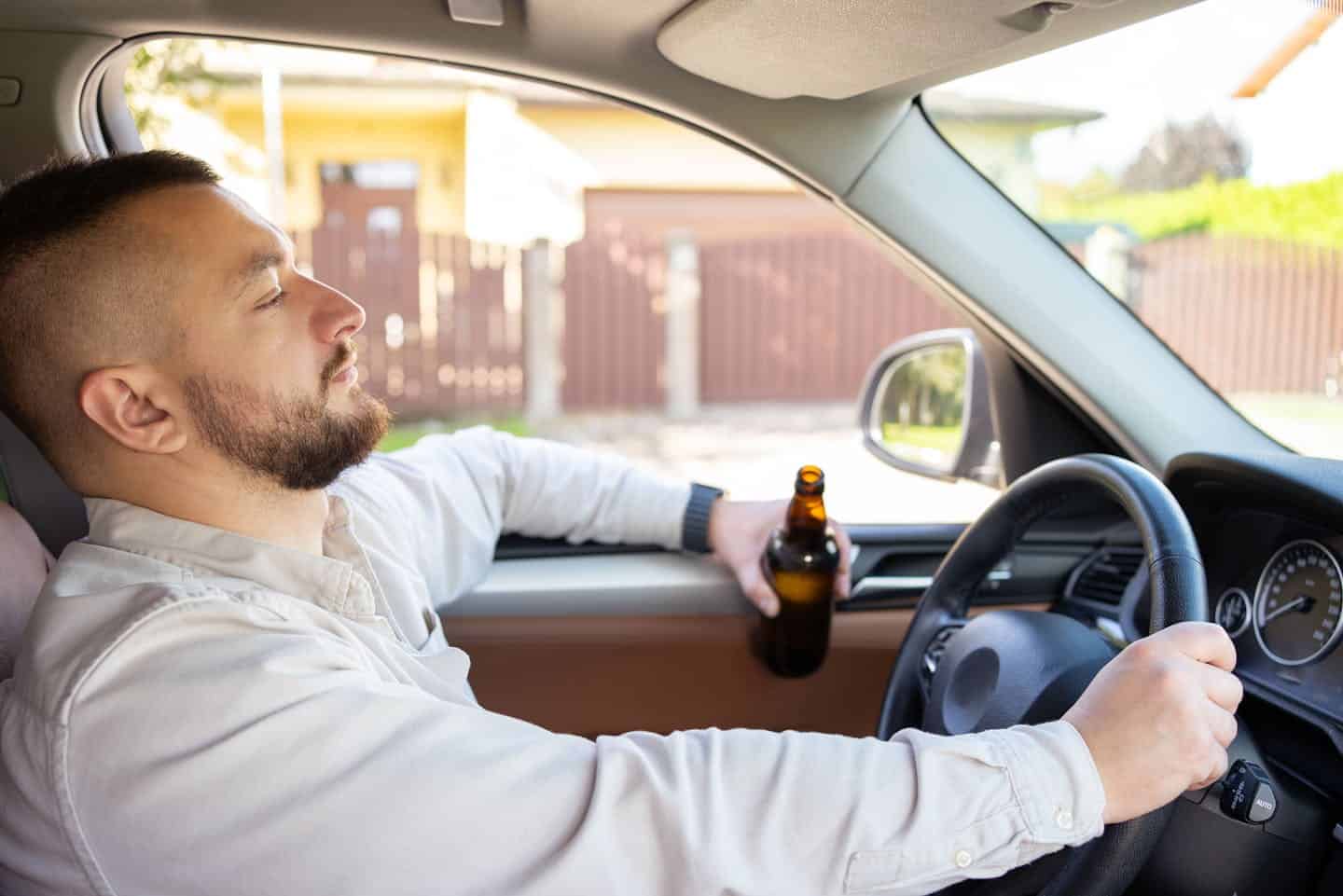 A man in a white shirt is holding a beer bottle while seemingly driving a car, depicted in a relaxed posture with an irresponsible behavior, highlighting a dangerous and illegal action.