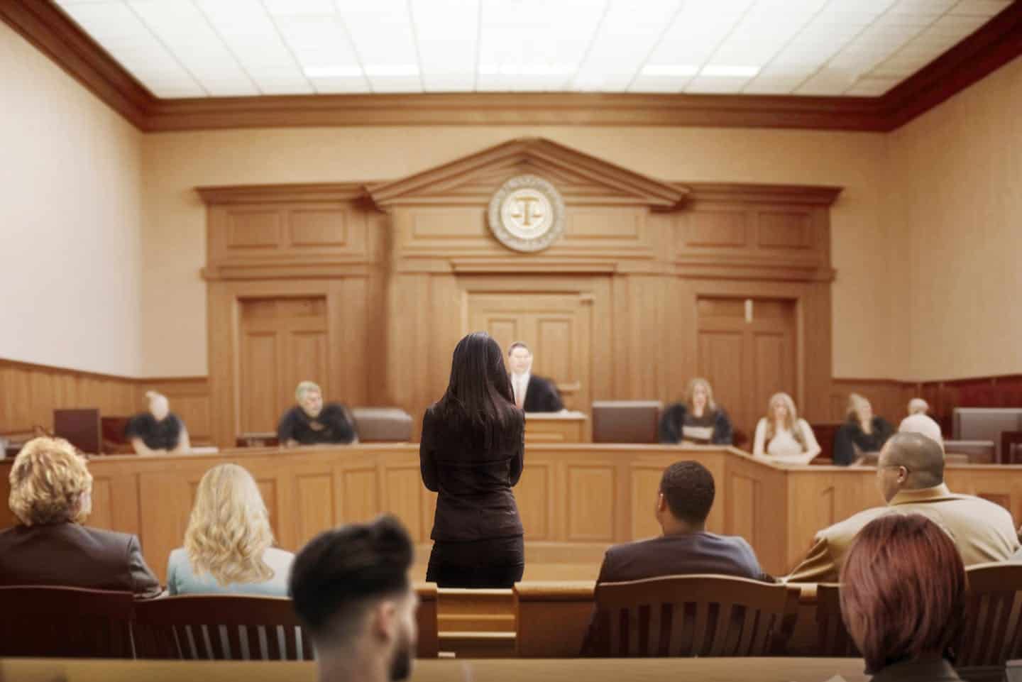 A woman stands addressing a courtroom, where a judge and jurors are seated; a legal setting with wood paneling and formal attire suggests a trial or hearing in progress.