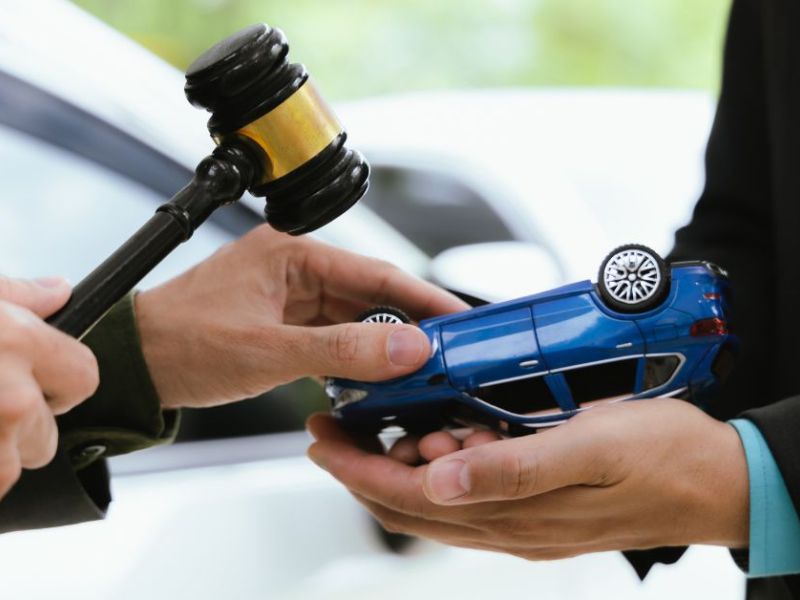 A person holding a gavel prepares to finalize a transaction, handing over a blue toy car to another individual, set in an outdoor environment with a blurred background of vehicles.